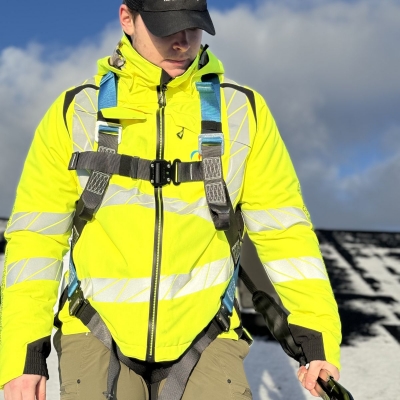 blå och svart fallskyddssele på man i varseljacka på tak med fallskyddsblock i handen