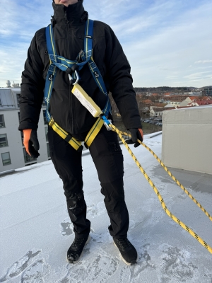 Man på tak i gul och blå fallskyddssele och gult rep med snö på taket